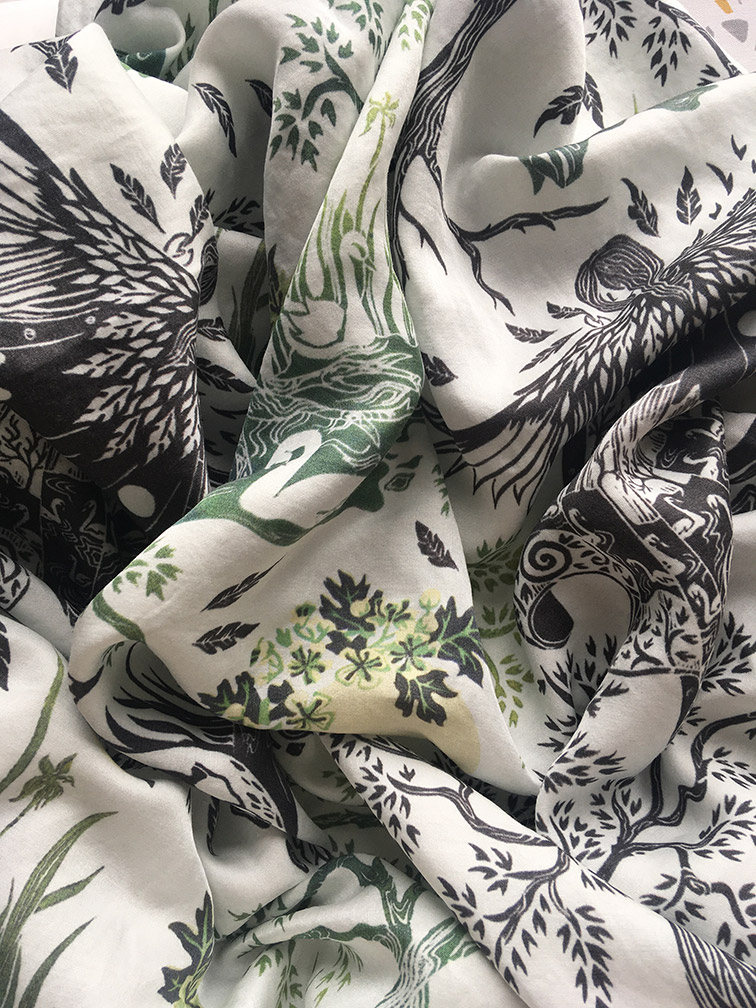 Photo of fabric with folds of material, the pattern on the fabric is partially hidden by the folds but includes leaves on branches, a moon with flowers, feathers, swans and the head and shoulders of a figure with wings