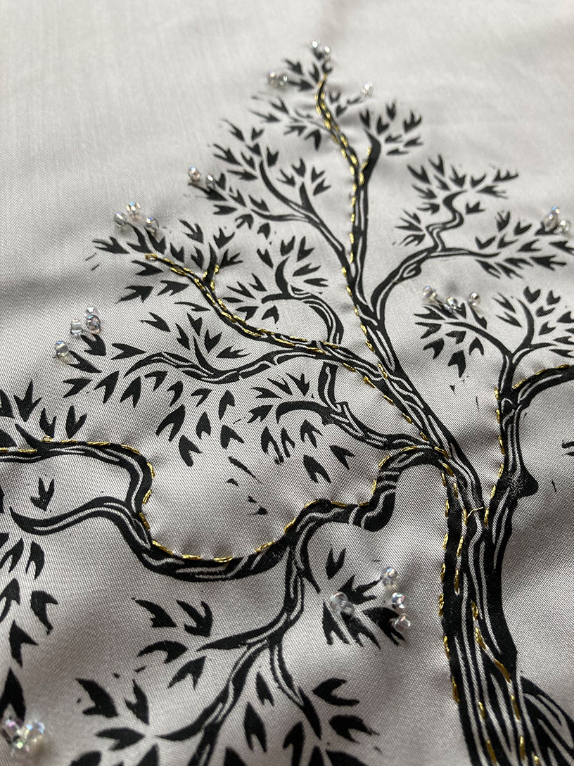 Photo of a black tree printed onto silver fabric with branches and leaves, these have some gold embroidery stitches on them and some clear glass beads.