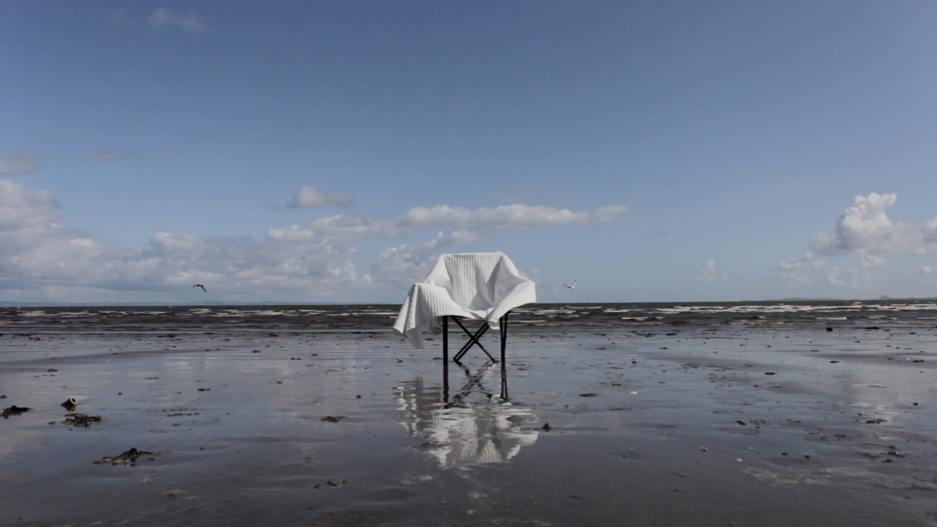 Wide angle shot of a chair on the shore line with a white blanket on top.