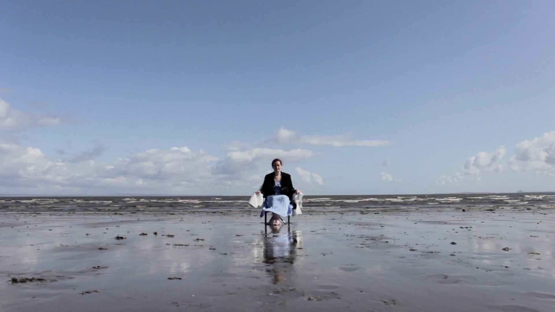A person sitting on a chair at the shoreline, the sandy beach in front of them and the sea behind. 