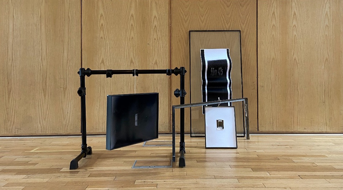 This image displays an installation shot of RESIDUES OF CUSTOMISATION in a wooden room. In this scenario, three art works positioned in a new formation, creating horizontal lines throughout the shot. The largest image held in the structure named ROTATIONS, ROTATIONS, ROTATIONS is largely indistinct, displaying a black expanse with a small, brighter outline in the centre of the image. Another structure in the background (STREAMY MINUTES) displays the sequence 55m 42s. 