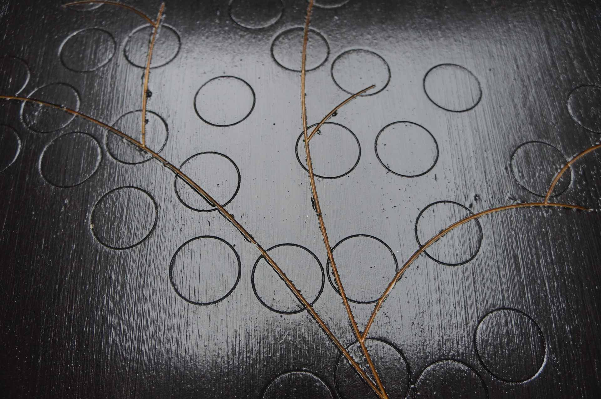 Close up photograph of a black painting, with about 15 neat circles and 3 lines carved into the wood.