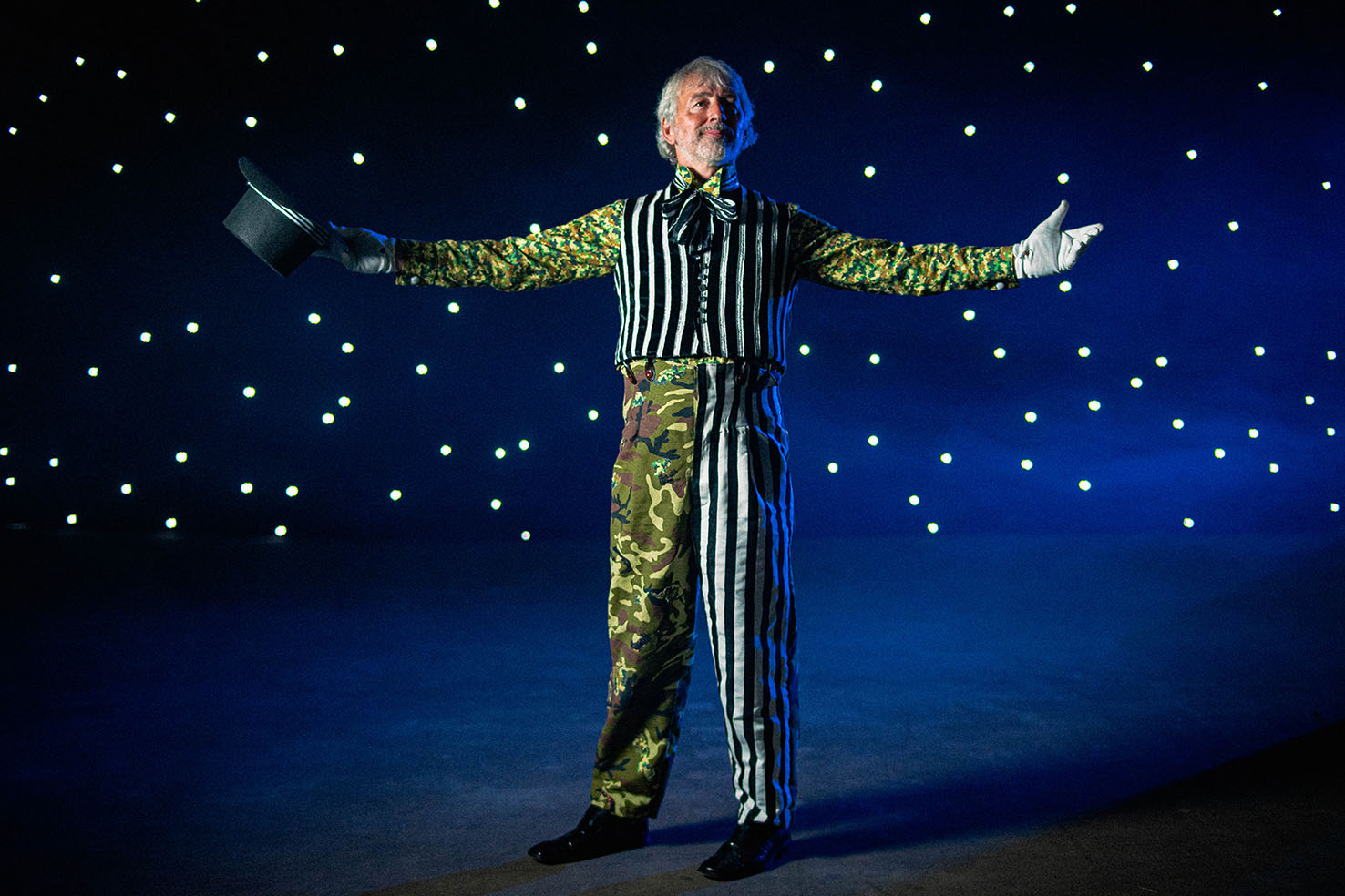 Prospero the Enchanter raising arms to the audience with a starry backdrop. He has removed his jacket and holds his top hat in his right hand.