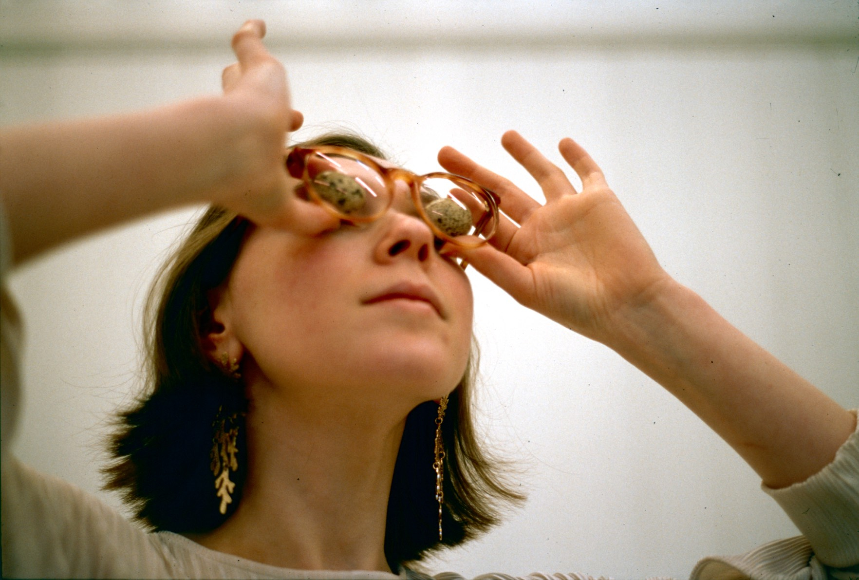 A photograph of a woman wearing glasses who is looking up whilst holding eggs up to her eyes