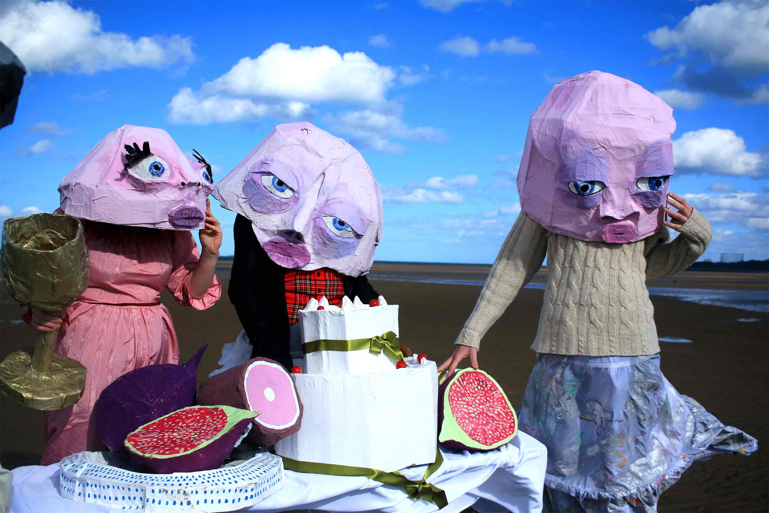 Three figures with pink papier-mâché masks stood around a table of cardboard food on the beach.