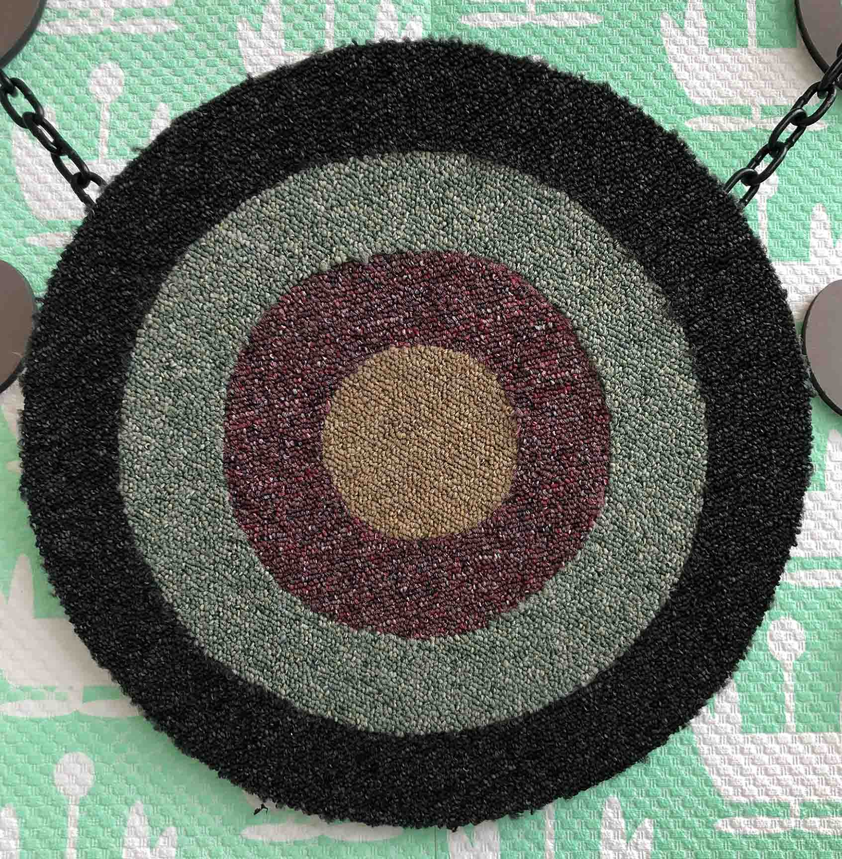 Target made from carpet, strung up with a black chain in front of a hand-printed pastel green wallpaper