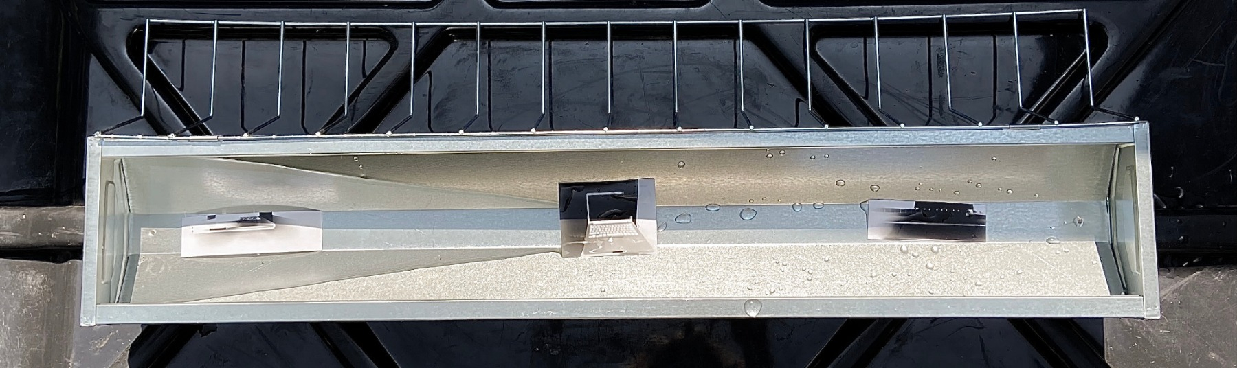 This image displays a metal trough that is filled with a shallow amount of water and carries three small photographs. The black and white photographs display images of technological devices such as laptops. They float within the water and appear wet.  