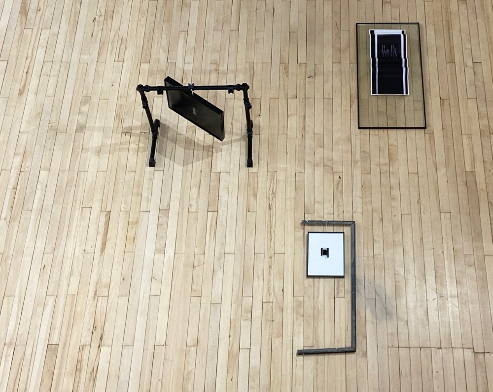 This image displays an installation shot of a flat lay view of RESIDUES OF CUSTOMISATION. In this scenario, three art works positioned in an organic formation on a wooden floor. The height that the image was taken at renders the art works slightly indistinct. The three art works appear to be made from linear structures, and display varying black and white photographic images. One image faintly reads the sequence 55m 42s. 